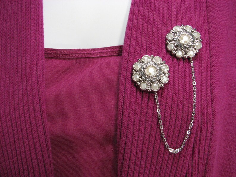 May include: Two silver-toned brooches with a chain connecting them. Each brooch features a circular design with white faux pearls and clear rhinestones.