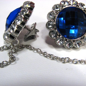 May include: A pair of silver cufflinks featuring a large, faceted, deep blue central stone, encircled by clear crystals. The cufflinks are linked by a fine chain.