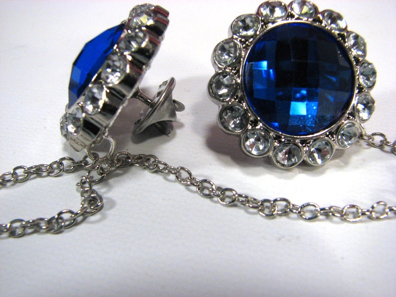May include: A pair of silver cufflinks featuring a large, faceted, deep blue central stone, encircled by clear crystals. The cufflinks are linked by a fine chain.
