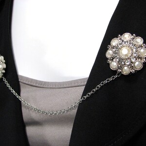 May include: A silver chain with two decorative brooches featuring white pearl-like stones and clear rhinestones. The brooches are connected by a silver chain.