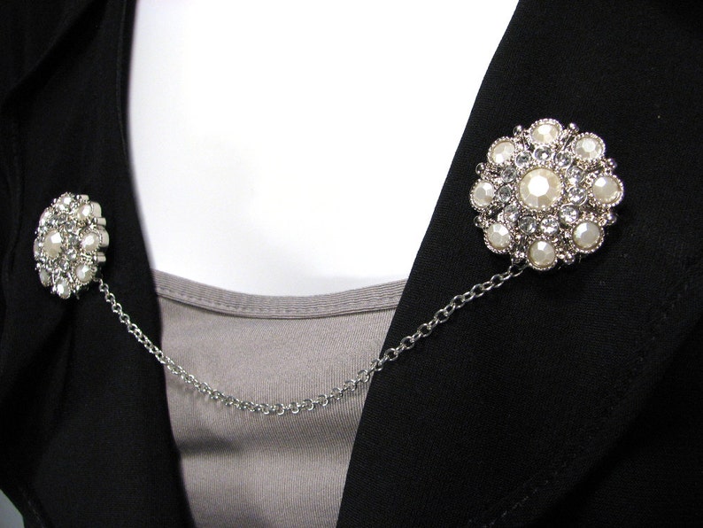 May include: A silver chain with two decorative brooches featuring white pearl-like stones and clear rhinestones. The brooches are connected by a silver chain.