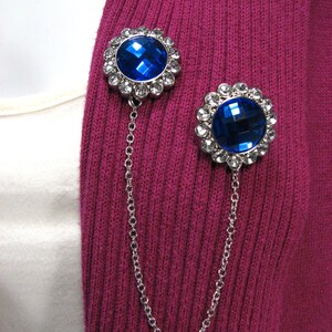 May include: Two brooches with large, faceted blue gemstones and a silver chain. The brooches are set against a ribbed, magenta fabric. Each brooch features a halo of smaller, clear crystals around the central blue stone. The chain connects the two brooches.
