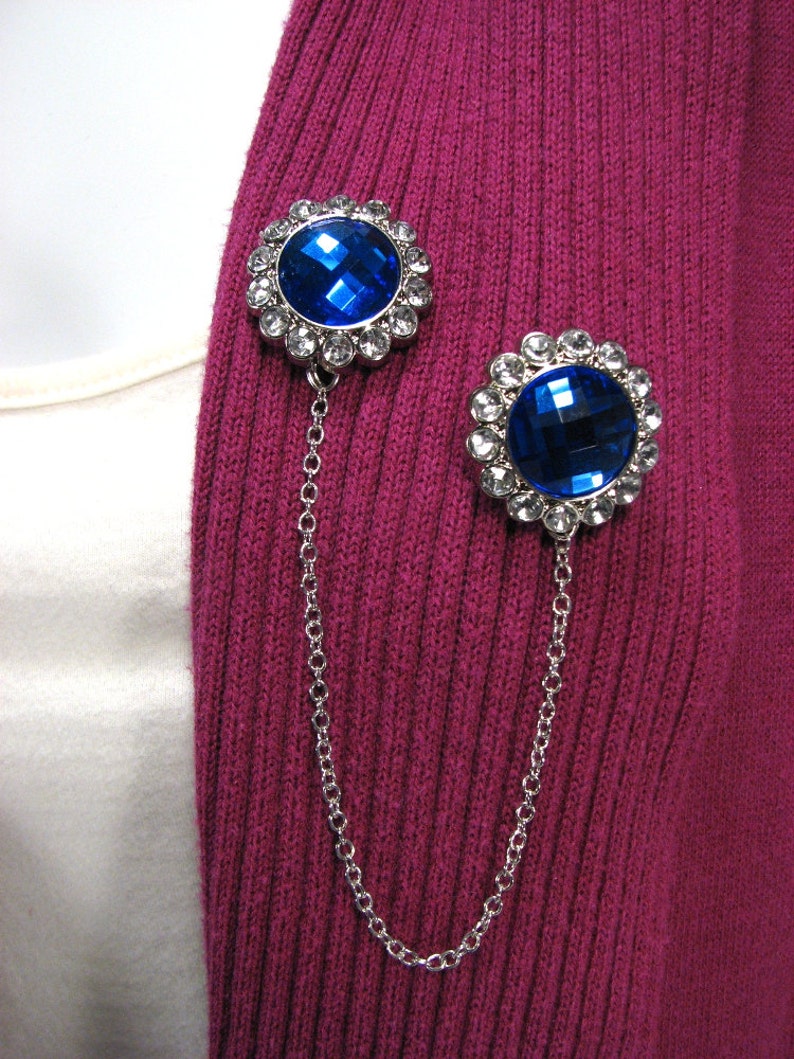 May include: Two brooches with large, faceted blue gemstones and a silver chain. The brooches are set against a ribbed, magenta fabric. Each brooch features a halo of smaller, clear crystals around the central blue stone. The chain connects the two brooches.