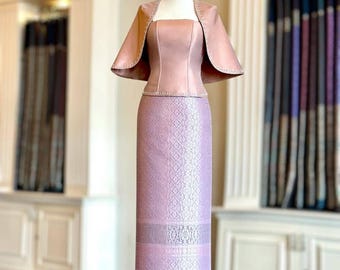 May include: A two-piece ensemble featuring a dusty rose sleeveless bodice with a square neckline and a matching capelet with stitched edges. The long, straight skirt is a light lavender hue with a detailed, repeating woven pattern throughout.
