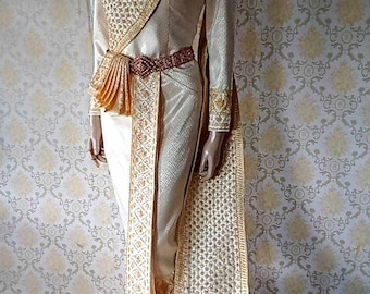 May include: A cream-colored long-sleeved dress featuring a structured high collar and a textured, gold-patterned sash draped diagonally across the torso. The garment includes a pleated waist detail, a central vertical panel, and a long train.