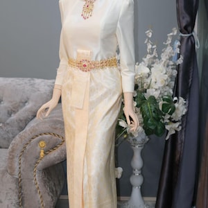 May include: A cream-colored long-sleeved blouse paired with a matching floor-length skirt featuring a subtle woven pattern. The outfit is accessorized with a gold-toned belt and necklace, both adorned with pink and clear stones, worn on a mannequin.