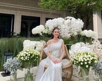 May include: A floor-length, one-shoulder silver gown with intricate embroidery and a fitted bodice, seated on a round, light-brown woven rattan chair. The dress features a draped neckline and a long, flowing skirt that pools on the ground.
