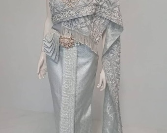 May include: A silver-toned traditional Thai ensemble featuring a one-shoulder bodice with intricate embroidery and a matching floor-length skirt. The outfit includes a decorative metallic belt with a central floral motif and a long, draped shawl.