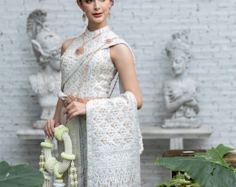 May include: A traditional Thai dress in white and silver featuring intricate embroidery and beadwork. The sleeveless bodice has a high neckline, paired with a matching wrap skirt and a draped shoulder sash. A floral garland is held in the hand.