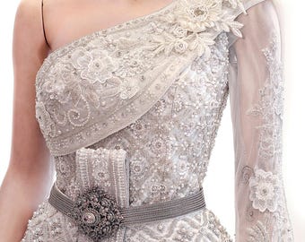 May include: An off-the-shoulder gown in a light silver hue, featuring intricate beadwork, sequins, and floral lace appliqués. The garment includes a high-neck collar, a long sheer sleeve, and a metallic belt with a large, ornate, dark circular buckle.