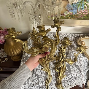 Puede incluir: Apliques de pared dorados y ornamentados con un diseño floral. Los apliques tienen una forma curva y decorativa y están hechos de metal. La imagen también muestra una lámpara de araña de cristal transparente con cristales colgantes y un jarrón dorado.