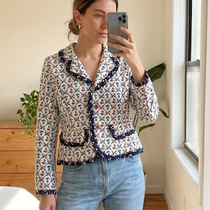 May include: A white blazer with a repeating anchor pattern in red and blue. The blazer has a navy blue trim and red buttons. The jacket is paired with light blue jeans.