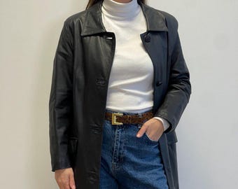 May include: A black leather coat with a classic collar and button closure. The coat is worn over a white turtleneck and blue jeans. A brown belt with a gold buckle adds a touch of detail to the outfit. The coat appears to be knee-length.
