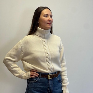 May include: A white cable knit turtleneck sweater with long sleeves and a brown leather belt. The sweater is worn over blue jeans.