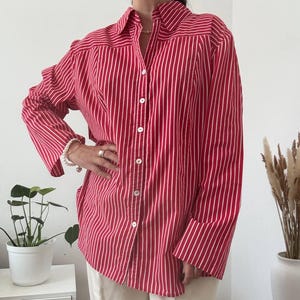 May include: A long-sleeved button-down shirt featuring a vibrant red and white vertical striped pattern. The shirt has a classic collar, a full-length front button placket with white buttons, and loose-fitting sleeves, paired with plain white trousers.