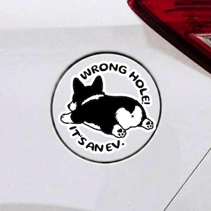 May include: A circular white sticker featuring a black and white illustration of a Corgi lying down, viewed from behind. The text WRONG HOLE! IT'S AN EV! is printed in black capital letters in a circular arc around the dog illustration.