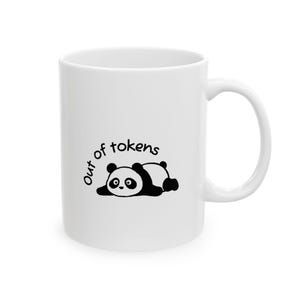 May include: A white ceramic mug featuring a black graphic of a panda lying down on its stomach. Above the panda, the text "Out of tokens" is printed in a simple black sans-serif font. The mug has a standard cylindrical shape with a side handle.