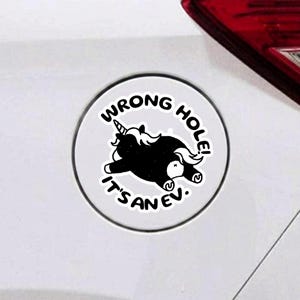 May include: A circular white sticker featuring a black graphic of a unicorn lying down. The text reads WRONG HOLE! IT'S AN EV. The sticker is applied to a white surface near a red vehicle taillight, displaying a humorous design for electric vehicles.