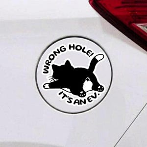 May include: A circular white sticker featuring a black and white illustration of a cat lying down with its rear end facing forward. The text WRONG HOLE! IT'S AN EV. is printed in black capital letters in an arc around the cat on the white background.