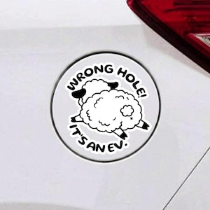 May include: A circular white sticker featuring a black and white cartoon illustration of a fluffy sheep. The text WRONG HOLE! IT'S AN EV! is printed in bold black capital letters around the sheep, which is positioned on a white surface.