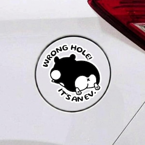 May include: A circular sticker featuring a black and white illustration of a hamster viewed from behind. The text WRONG HOLE! IT'S AN EV. is printed in black capital letters around the hamster. The sticker is applied to a white surface.