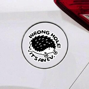 May include: A circular white sticker featuring a black and white illustration of a hedgehog. The text reads WRONG HOLE! IT'S AN EV. The sticker is applied to a white surface near a red vehicle taillight, displaying a humorous graphic design.