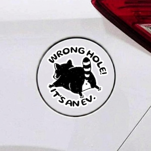 May include: A circular white sticker featuring a black silhouette of a raccoon facing away with its tail raised. The text WRONG HOLE! IT'S AN EV. is printed in black capital letters around the raccoon graphic on the sticker surface.