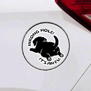 May include: A circular white sticker featuring a black silhouette of a puppy lying down with its rear end facing forward. The text WRONG HOLE! IT'S AN EV. is printed in black capital letters in a curved arc around the puppy illustration.