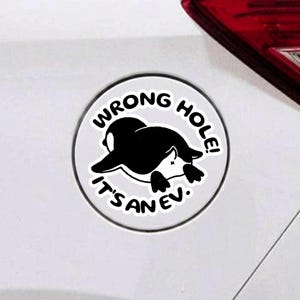 May include: A circular white sticker with a black border featuring a black and white illustration of a penguin lying face down. The text WRONG HOLE! IT'S AN EV. is printed in black capital letters in a circular arc around the penguin graphic.