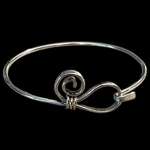 May include: Silver-toned bangle bracelet with a decorative swirl and hook closure. The bracelet features a spiral design and a simple, elegant aesthetic. The jewelry is set against a black background.