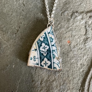 May include: A silver necklace featuring a triangular pendant. The pendant is a fragment of ceramic with a teal and white floral pattern, encased in a silver frame. The chain is a delicate silver link.