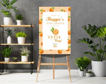 May include: A welcome sign for a baby shower with an orange theme. The sign is on an easel and has a white background with orange slices and the text "Welcome to Maggie's Baby Shower Celebrating a Little Cutie on the Way! August 20".