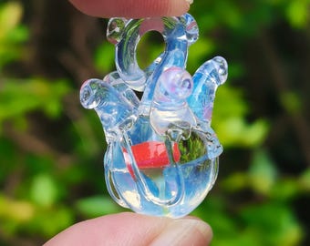May include: A clear glass pendant shaped like an anatomical heart with iridescent blue and white highlights. The pendant features a small red accent inside the glass structure and a circular loop at the top for hanging as a necklace charm.