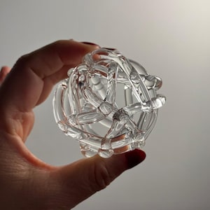 May include: A clear, spherical glass object with an intricate, interwoven design. The transparent orb is held in a hand, showcasing its complex, geometric structure. The light reflects off the glass, highlighting its curves and details.