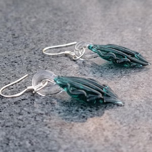 Peut inclure: Une paire de boucles d'oreilles en argent avec des pendentifs en verre bleu sarcelle en forme de larme. Le verre est texturé et présente un motif tourbillonnant.