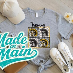May include: A light gray t-shirt with a graphic of Trojan helmets in a grid pattern. The word "Trojans" is above the graphic. A white baseball cap and white sneakers are also in the image. The words "Made on Main" are in a teal script.