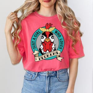 May include: A coral-colored t-shirt with a graphic of a cartoon chicken wearing a yellow polka-dot bow. The text reads "Just a girl who loves peckers" in a circular design.