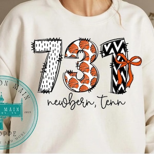 May include: Cream-colored sweatshirt with the numbers "731" in a graphic design. The "7" is black and white, the "3" has a pumpkin pattern, and the "1" is black and white with an orange bow. The text "newbern, tenn" is below.