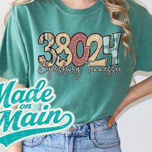 May include: Teal green t-shirt with a graphic design featuring the numbers "38024" in various patterns and colors, with "Dyersburg, Tennessee" below. The shirt also has the text "Made on Main" in a teal script.