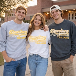May include: Three people wearing sweatshirts with the text "Dyersburg Trojans" in yellow and white. The sweatshirts are gray, white, and dark gray. The people are smiling and standing outside a brick building.