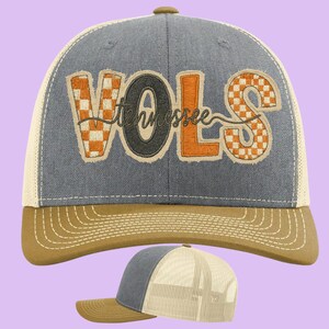 May include: A trucker hat with a gray front panel, tan brim, and a white mesh back. The hat features the word "VOLS" in orange and checkered orange and white, with "Tennessee" embroidered in a script font.