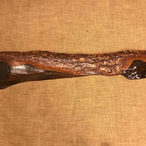Op de afbeelding: Een lang, dun stuk hout met een natuurlijke, rustieke afwerking. Het hout heeft een donkerbruine kleur met lichtere bruine en roodbruine strepen. Het hout is glad en gepolijst.