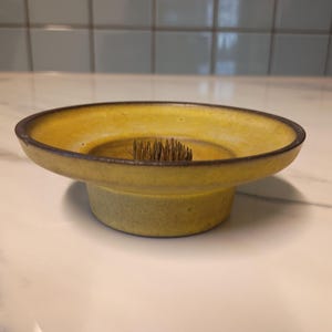 Peut inclure: Un bol pique-fleurs circulaire en céramique jaune avec un bord brun foncé. Le bol présente un support à épingles en métal central avec de nombreuses pointes verticales conçues pour arranger les fleurs, fixé sur une base jaune à large bord.