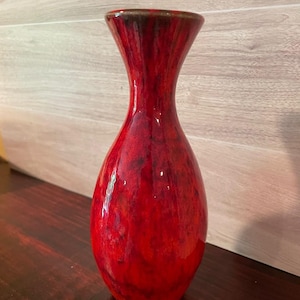 Może przedstawiać: Wysoki, błyszczący, czerwony wazon z rozszerzaną górą i zaokrąglonym korpusem. Wazon ma ciemnobrązową obwódkę i teksturowany, nakrapiany wygląd. Wazon jest prawdopodobnie ceramiczny lub szklany i nadaje się do eksponowania kwiatów lub jako przedmiot dekoracyjny.