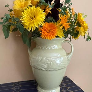 May include: A cream-colored ceramic pitcher with two handles and a decorative embossed leaf pattern around the center, filled with a vibrant bouquet of yellow and orange chrysanthemums, daylilies, and dark purple pansies with green foliage.
