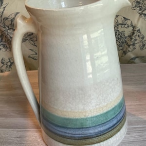 May include: Cream-colored ceramic pitcher with a handle and spout. The pitcher features horizontal bands of blue, green, and tan near the base. The surface has a crackle glaze effect, adding texture to the piece. A decorative kitchenware item.