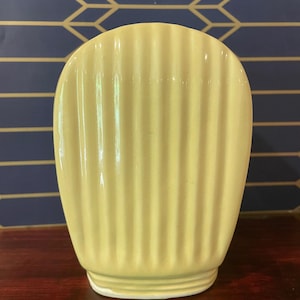 May include: A pale yellow, ribbed ceramic vase with a rounded top and a slightly flared base. The vase has vertical ridges and a smooth, glossy finish. The background is a dark blue wall with a gold geometric pattern.