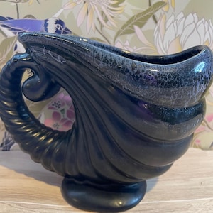 May include: A black ceramic vase shaped like a seashell with a curled handle. The vase has a speckled glaze.