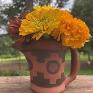 May include: A small, terracotta-colored pitcher with a handle and a decorative gear pattern. The pitcher is filled with a vibrant arrangement of yellow and orange flowers, with a few red blooms. The pitcher is sitting on a wooden surface.