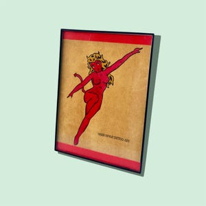 May include: Framed artwork featuring a red devil woman with yellow hair and a tail, set against a tan background. The artwork is framed in a black frame with a red border. The text "TIGER STYLE TATTOO ART" is printed at the bottom.